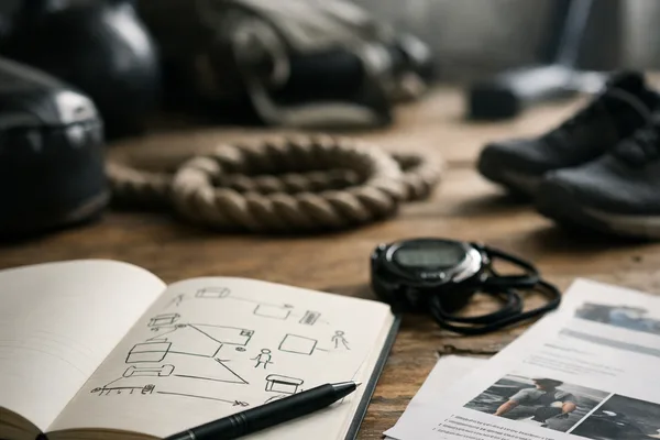 Aufgeschlagenes Notizbuch mit Skizzen, Stift, Stoppuhr und Ausdrucken auf Holztisch, Trainingsgeräte unscharf