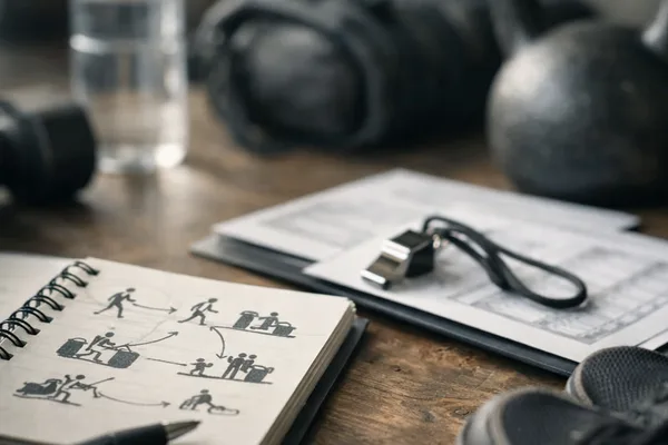 Notizbuch mit Trainingsskizzen, Trillerpfeife, Kettlebell, Wasserflasche, Sporttasche und Schuhen auf Holztisch