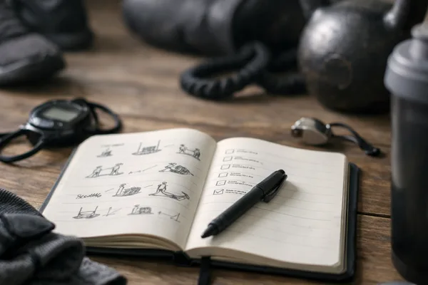 Aufgeschlagenes Notizbuch mit Skizzen und Stift auf Holztisch, dahinter Kettlebell, Stoppuhr, Schuhe