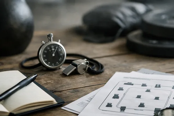 Stoppuhr, Trillerpfeife, Notizbuch mit Stift und Trainingsplan auf Holztisch, Kettlebell im Hintergrund