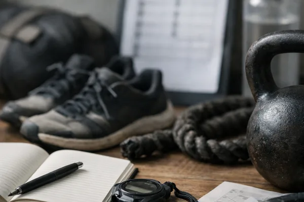 Fitnessutensilien auf Holztisch: Notizbuch, Stoppuhr, Kettlebell, Laufschuhe, Tau, Klemmbrett und Trinkflasche