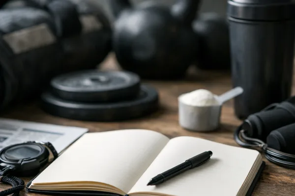 Leeres Notizbuch mit Stift auf Holztisch, dahinter Kettlebells, Shaker, Messlöffel mit Pulver